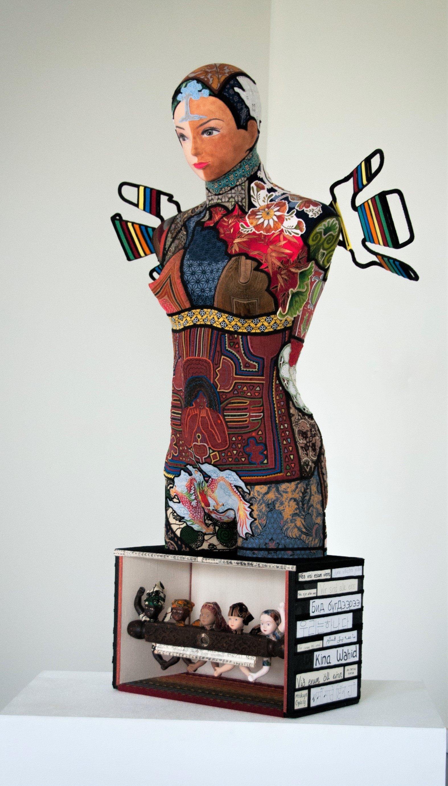 "We are One" sculpture by Libby Bloxham. A mannequin of a woman is covered in a patchwork of recycled textiles suggesting different cultures and styles. The mannequin lacks arms and legs but has wings also formed from the textiles. The base of the sculpture is a box containing the upper bodies of various small dolls depicting a spectrum of ethnicities.