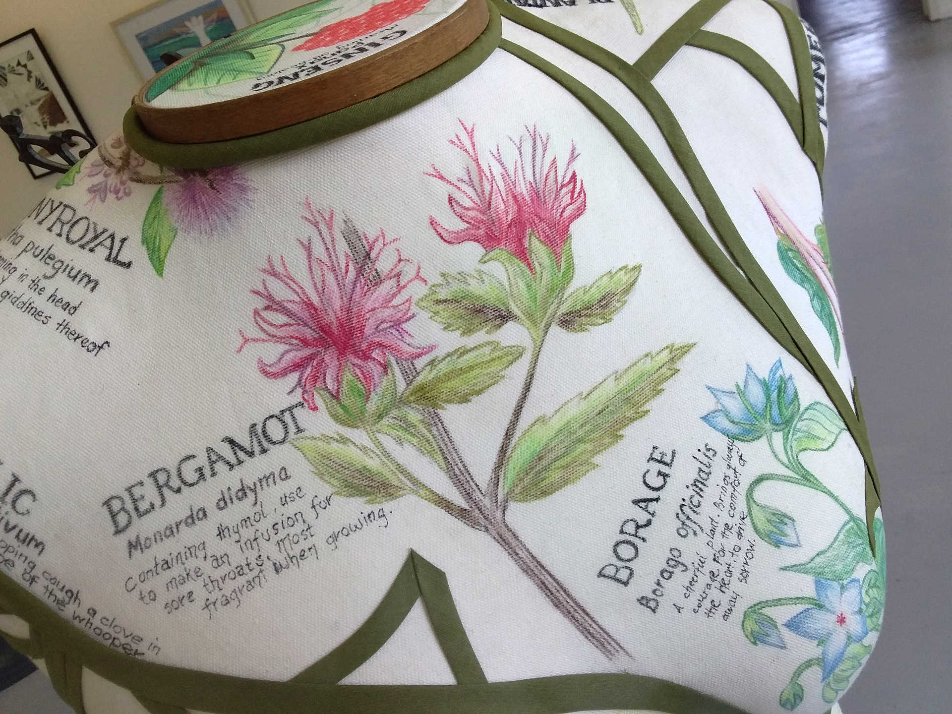 A close-up detail shot of 'Disempowerment' by Libby Bloxham, showing a white fabric panel covering the chest of a female torso mannequin. The panel contains various pencil illustrations of botanical specimens, with two clearly visible from this angle. One is a branch with jagged green leaves and sprawling red/pink flowers, and the text: 'Bergamot', 'Monarda didyma', 'Containing thymol, use to make an infusion for sore throats. Most fragrant when growing.'. The second is bright blue flower with smooth leaves, and the text: 'Borage', 'Borago officinalis', 'A cheerful plant. Brings always courage. For the comfort of the heart, to drive away sorrow.'. 