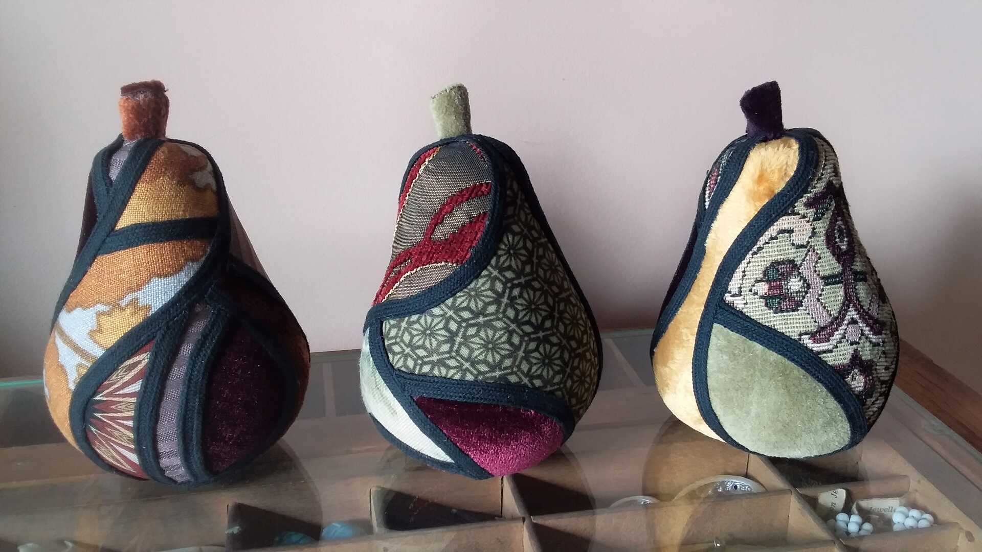 Sculpture 'Enough for Everyone II' by Libby Bloxham. Three pears are covered in swatches of fabric, in shades favouring olive green, orange and red. The fabric covering is segmented into irregular panels with thick black binding, complimenting the contours of the shape.