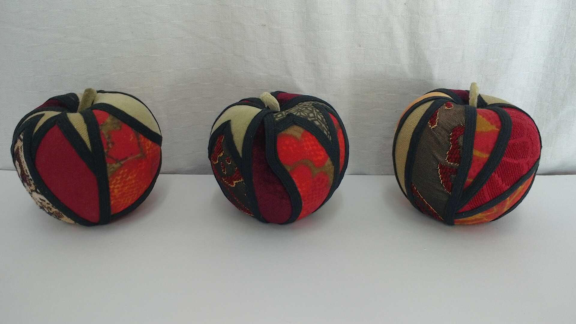 Sculpture 'Enough for Everyone I' by Libby Bloxham. Three Apples are covered in swatches of fabric in shades of olive green and red. The fabric covering is segmented into irregular panels with thick black binding, complimenting the contours of the shape.