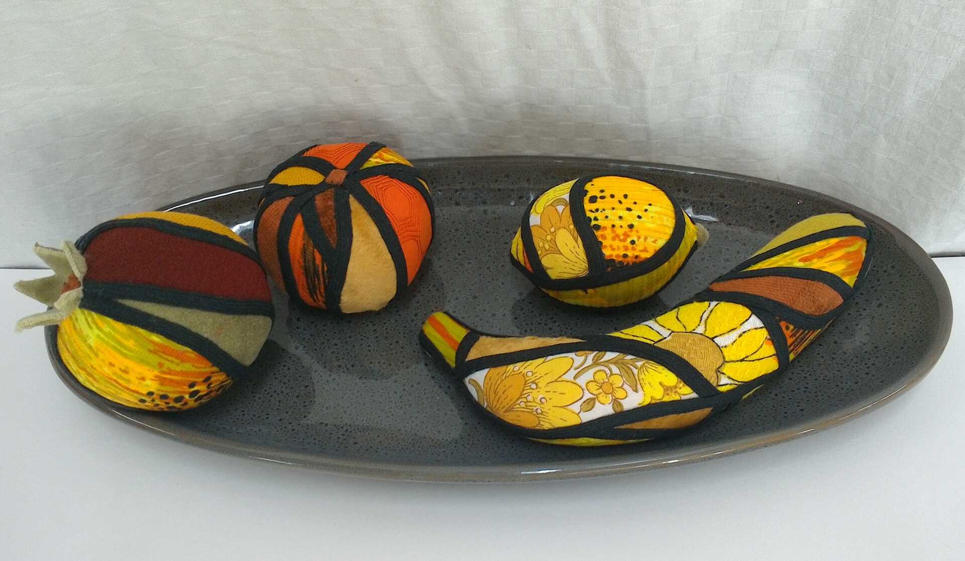 Sculpture 'The Fruit Bowl' by Libby Bloxham. Arranged on an oval platter are four pieces of fruit - including a banana, lemon and orange - covered in swatches of fabric favouring yellow, orange and red tones. The fabric covering is segmented into irregular panels with thick black binding, complimenting the contours of the shape.