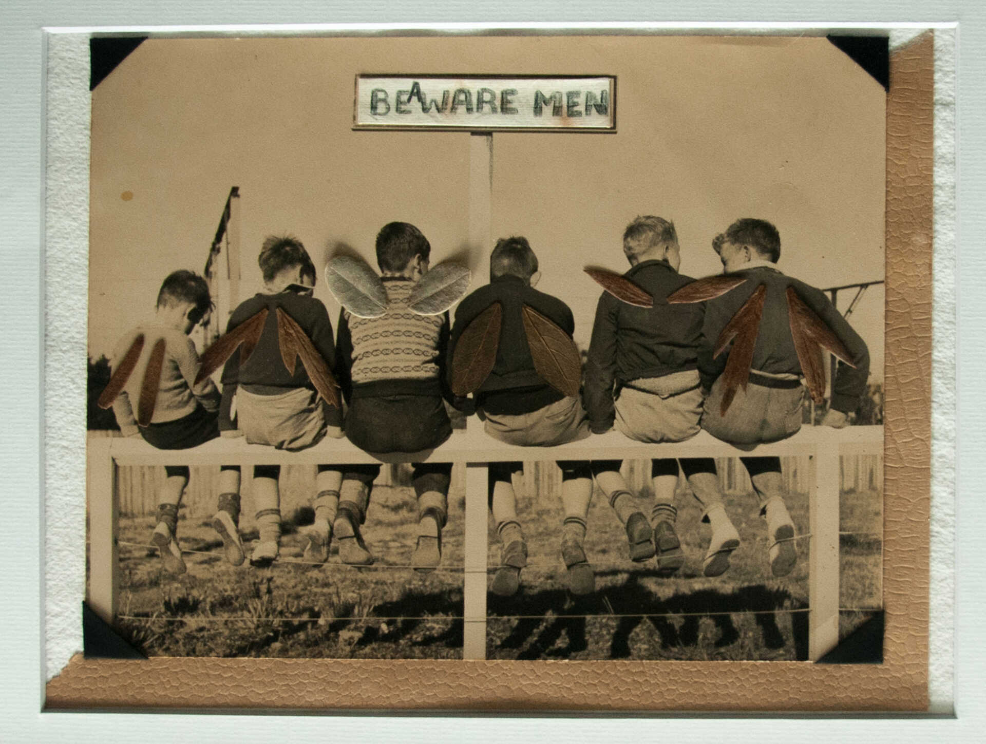 Collage work "Aware Men I" by Libby Bloxham. A sepia-toned vintage photograph shows a row of children—apparently young boys—seated on a fence, seen from behind. Small leaves are positioned at the shoulders of each figure, in pairs, so they look like wings. in the background, facing the figures and the viewer, is a sign saying "Beware Men", two which an additional "A" has been added to instead make "Be Aware Men".