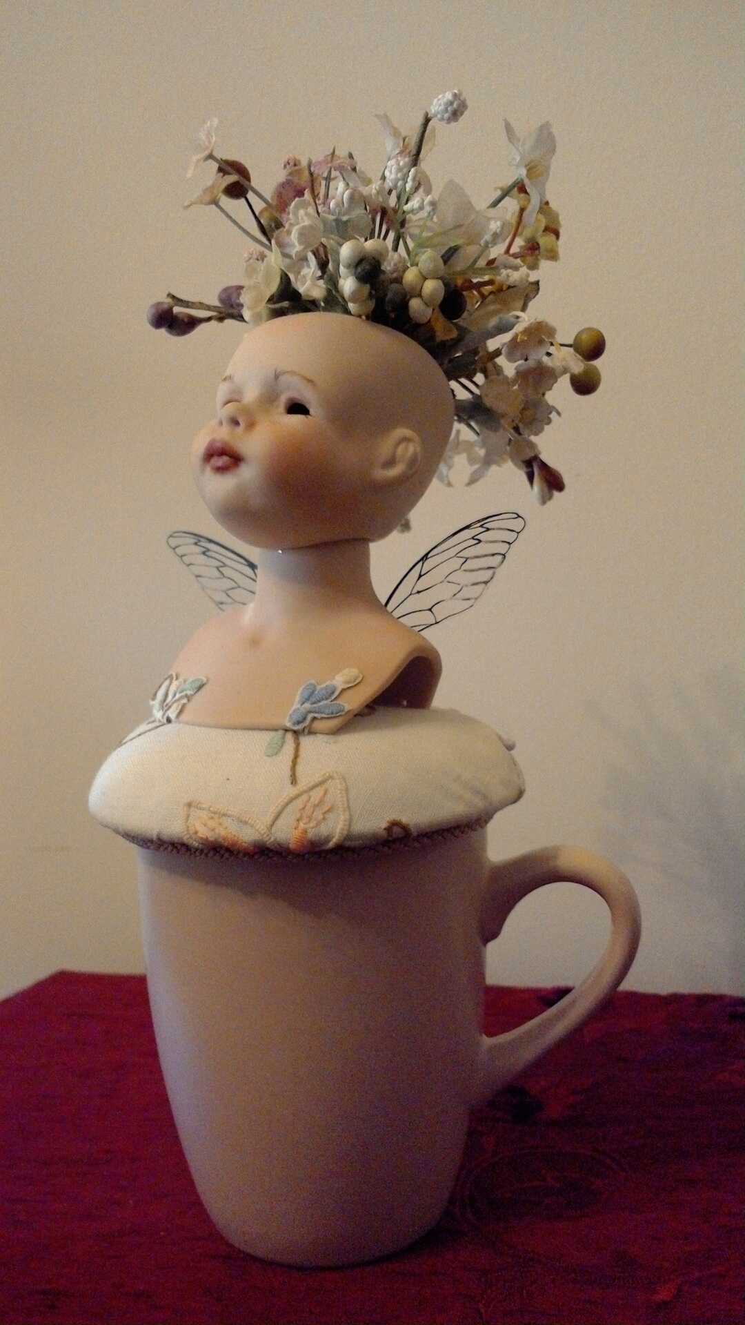 Sculpture "Finding the Way Back III" by Libby Bloxham. The head and shoulders of a porcelain doll are posed atop a rose pink drinking mug, on a round white fabric cushion with an embroidered flower pattern. The doll has no hair affixed, but a large cluster of flowers sprout wildly from her crown. Her head is raised with a proud expression, and acetate insect wings rise from her back.