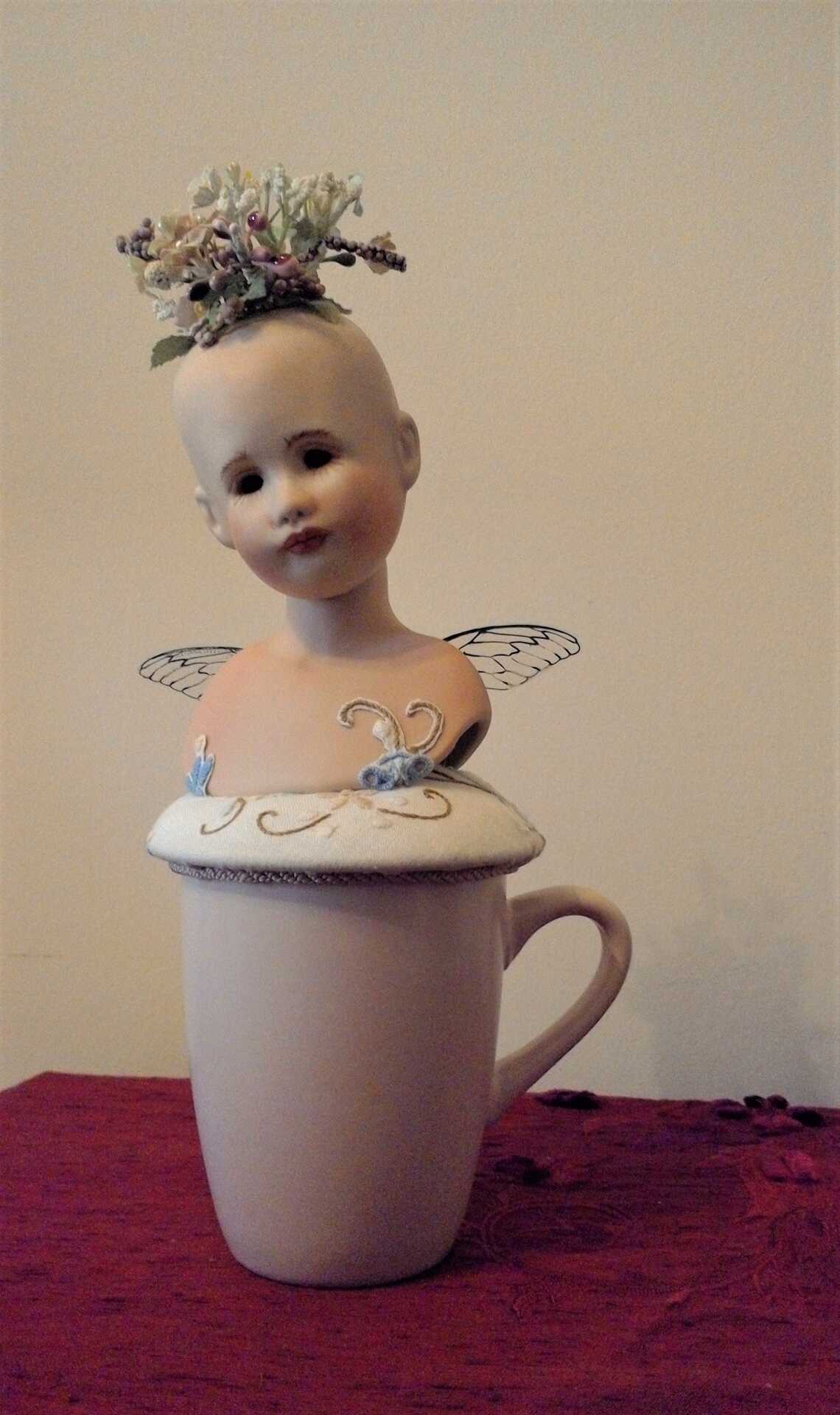 Sculpture "Finding the Way Back II" by Libby Bloxham. The head and shoulders of a porcelain doll are posed atop a rose pink drinking mug, on a round white fabric cushion with an embroidered flower pattern. The doll has no hair affixed, but a cluster of flowers sprout from her crown. Her head is tilted to one side with a wistful expression, and acetate insect wings droop slightly at her back.