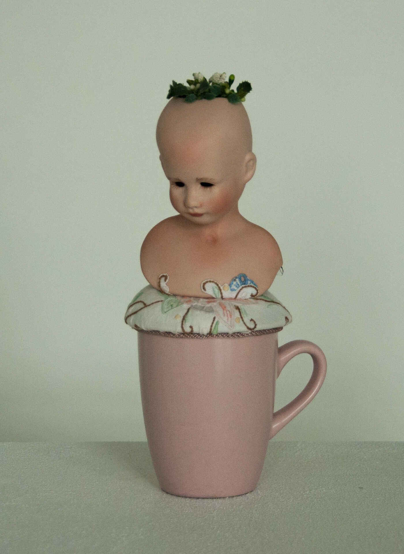 Sculpture "Finding the Way Back I" by Libby Bloxham. The head and shoulders of a porcelain doll are posed atop a rose pink drinking mug, on a round white fabric cushion with an embroidered flower pattern. The doll has no hair affixed, but a small cluster of flower buds sprout from her crown. Her pose is demure, her eyes cast slightly downward; from the angle she is photographed you can barely see a tiny pair of acetate insect wings growing from her back.