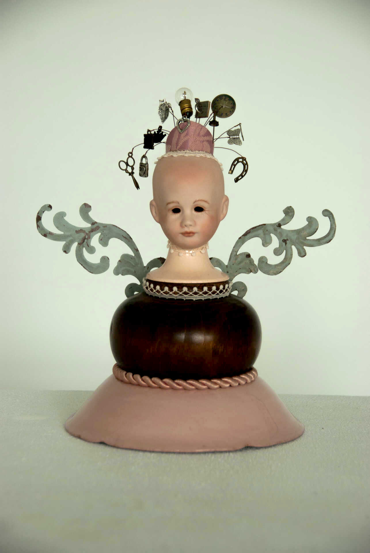 Sculpture "Gather the Tools" by Libby Bloxham. The head and shoulders of a porcelain doll are posed atop a wooden bulb and rose pink base, decorated with white lace and pink brocade. The doll has no hair affixed, but wears a small, rose pink cap. Suspended from the cap, on gently curving wires, are all manner of tools and trinkets, including scissors, a horseshoe, a padlock and a lightbulb. Metal flourishes, art nouveau style with a weathered grey-green finish, suggest the shape of wings where the doll's neck meets the sculpture's base.