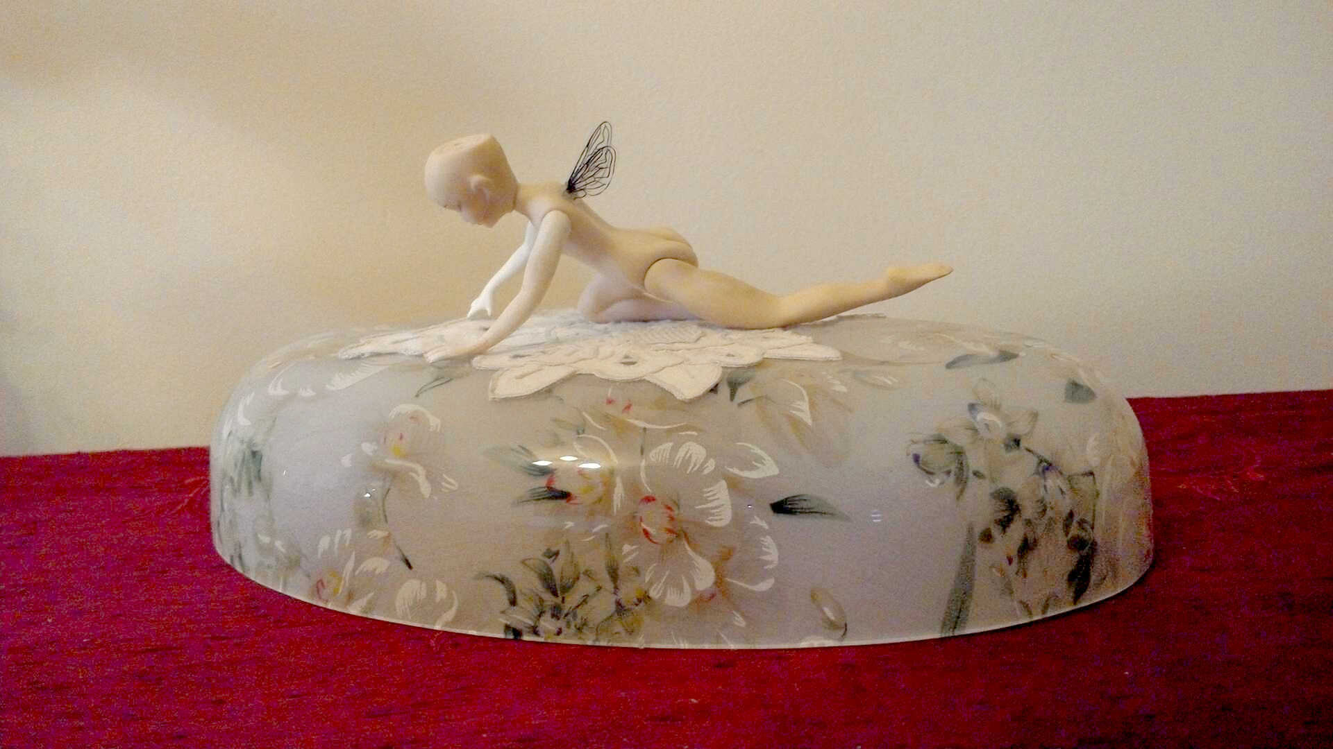 Sculpture "In the Face of Reality" by Libby Bloxham. A small articulated doll, with no hair or clothing, attached, is posed on top of a glass dome painted with floral designs, beneath her a white lace doily. The doll is facing downward, arms extended before her, kneeling on one leg with the other outstretched behind, giving the impression she is either landing or rising from where she rests. Acetate insect wings, very small compared to the doll's body, extend from her shoulders.