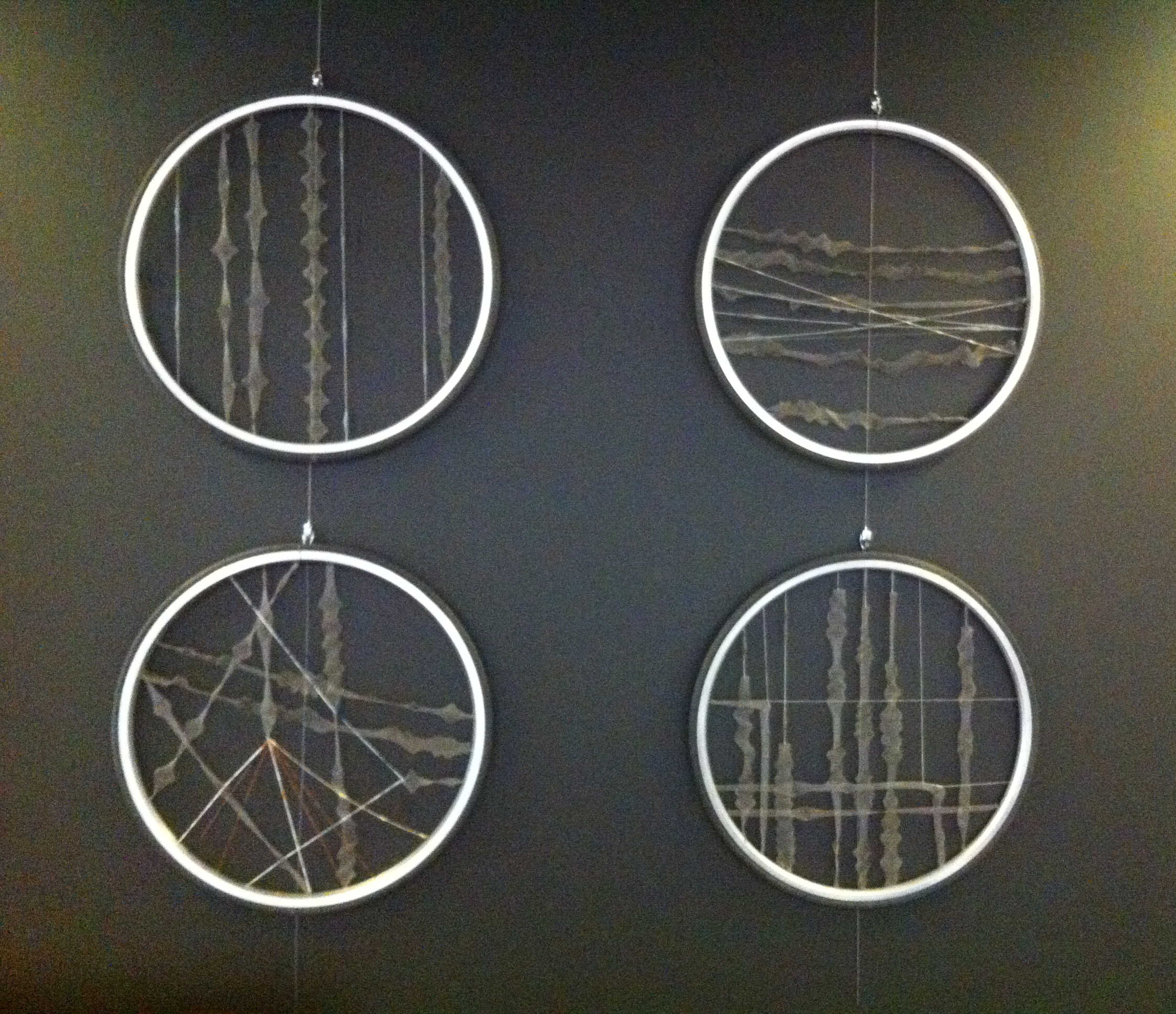 Sculpture "My Line of Site" by Libby Bloxham. Hanging in a 2x2 grid are four large hoops that appear to be bicycle tyres. Across the centre of each hoop are arranged many thin strips of floaty, irregularly shaped and gathered fabric, giving the impression of many straight lines. The first hoop features vertical lines, the second horizontal lines, the fourth a combination of vertical and horizontal lines, and the third diagonal lines in all directions.